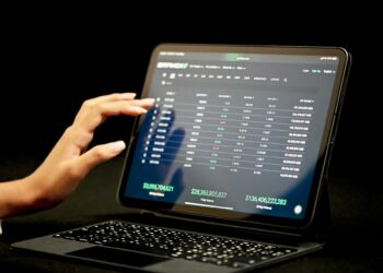 A person interacts with a financial trading platform on a touchscreen tablet, displaying various stock market data including numbers in green and red, and columns labeled with figures such as $22,365,605,532 and $746,406,223,869. The background is black, emphasizing their investment style. | 5StarsStocks