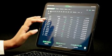 A person interacts with a financial trading platform on a touchscreen tablet, displaying various stock market data including numbers in green and red, and columns labeled with figures such as $22,365,605,532 and $746,406,223,869. The background is black, emphasizing their investment style. | 5StarsStocks