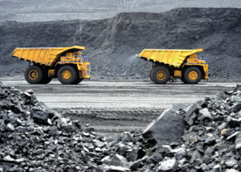 Two large yellow dump trucks, symbols of basic materials stocks, are visible in an open-pit mine. The trucks are positioned on a dirt path and face opposite directions. The foreground is filled with loose rocks, and the background shows the walls of the mine with layers of exposed earth. | 5StarsStocks
