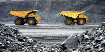 Two large yellow dump trucks, symbols of basic materials stocks, are visible in an open-pit mine. The trucks are positioned on a dirt path and face opposite directions. The foreground is filled with loose rocks, and the background shows the walls of the mine with layers of exposed earth. | 5StarsStocks
