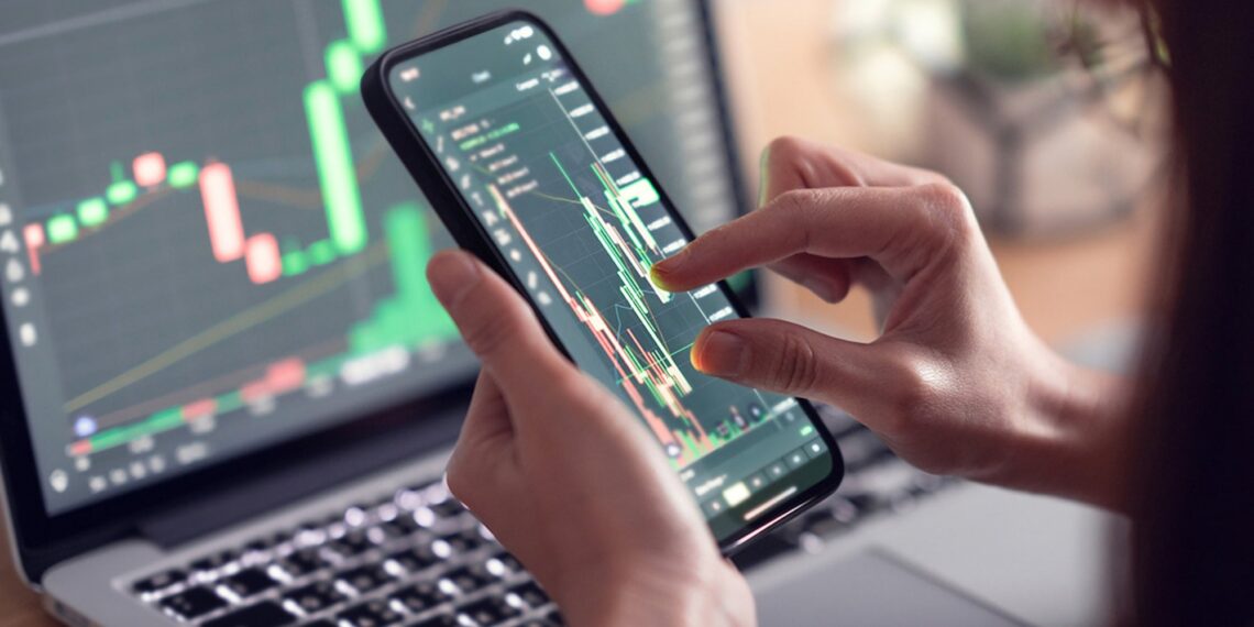 Close-up of a person holding a smartphone with a stock trading app displaying a candlestick chart. A laptop with similar financial charts is open in the background, suggesting active monitoring of stock market or cryptocurrency prices, possibly for credit repair strategies. | 5StarsStocks