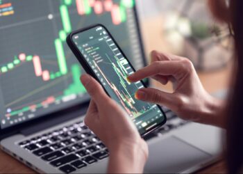 Close-up of a person holding a smartphone with a stock trading app displaying a candlestick chart. A laptop with similar financial charts is open in the background, suggesting active monitoring of stock market or cryptocurrency prices, possibly for credit repair strategies. | 5StarsStocks