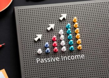 A vertically oriented perforated board displays a graph with colorful pegs forming upward-trending bars, resembling passive stocks growth. Three white arrows above the bars point upwards, indicating growth. The text "Passive Income" is written below the chart. Office supplies lie nearby. | 5StarsStocks