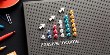 A vertically oriented perforated board displays a graph with colorful pegs forming upward-trending bars, resembling passive stocks growth. Three white arrows above the bars point upwards, indicating growth. The text "Passive Income" is written below the chart. Office supplies lie nearby. | 5StarsStocks