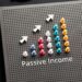 A vertically oriented perforated board displays a graph with colorful pegs forming upward-trending bars, resembling passive stocks growth. Three white arrows above the bars point upwards, indicating growth. The text "Passive Income" is written below the chart. Office supplies lie nearby. | 5StarsStocks