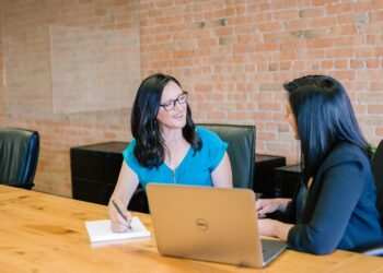 Two women are seated at a wooden table in an office setting. One is taking notes on a notepad while holding a pen, and the other has a laptop open in front of her. They are both smiling and appear to be engaged in a friendly conversation about hiring a financial advisor. | 5StarsStocks