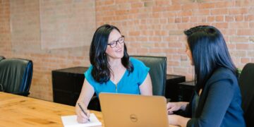 Two women are seated at a wooden table in an office setting. One is taking notes on a notepad while holding a pen, and the other has a laptop open in front of her. They are both smiling and appear to be engaged in a friendly conversation about hiring a financial advisor. | 5StarsStocks