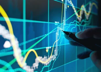 A hand points a pen at a digital screen displaying financial graphs and charts. Various colorful lines depict stock market trends, with bar graphs representing data points, including undervalued stocks. The background is dark with light blue and green neon-like hues highlighting the data. | 5StarsStocks