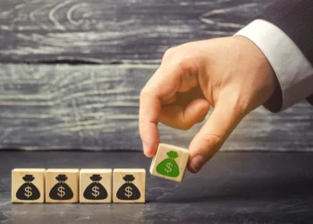 A hand is arranging wooden blocks on a table. Four blocks, each with an icon of a black money bag and a dollar sign, are aligned in a row. The hand is placing a fifth block with a green money bag and a dollar sign next to them, symbolizing the growth potential of income stocks. The background is a textured gray surface. | 5StarsStocks