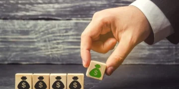 A hand is arranging wooden blocks on a table. Four blocks, each with an icon of a black money bag and a dollar sign, are aligned in a row. The hand is placing a fifth block with a green money bag and a dollar sign next to them, symbolizing the growth potential of income stocks. The background is a textured gray surface. | 5StarsStocks
