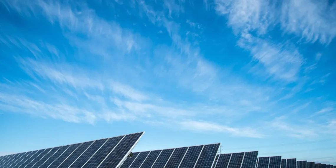 A wide array of solar panels installed on the ground under a clear blue sky with streaks of wispy clouds. The solar panels, which could impact energy stocks, are arranged in rows, capturing sunlight for renewable energy production. The landscape is flat, with dry grass between the panels. | 5StarsStocks