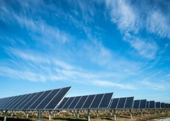 A wide array of solar panels installed on the ground under a clear blue sky with streaks of wispy clouds. The solar panels, which could impact energy stocks, are arranged in rows, capturing sunlight for renewable energy production. The landscape is flat, with dry grass between the panels. | 5StarsStocks