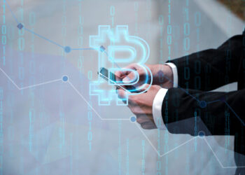 A person in a suit is holding a smartphone. A large, translucent Bitcoin symbol and binary code overlay the image, suggesting cryptocurrency trading or technology with 5StarsStocks.com Crypto. The background includes faint graphs and lines, indicating financial data or market analysis. | 5StarsStocks