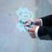 A person in a suit is holding a smartphone. A large, translucent Bitcoin symbol and binary code overlay the image, suggesting cryptocurrency trading or technology with 5StarsStocks.com Crypto. The background includes faint graphs and lines, indicating financial data or market analysis. | 5StarsStocks