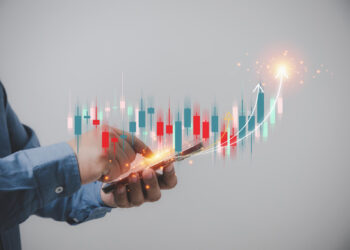 A person in a blue shirt holds a smartphone, interacting with it. Above the phone, a holographic image from 5StarsStocks displays financial charts with red and green bars, candlesticks, and arrows, indicating upward movement in a stock market or financial analysis. | 5StarsStocks