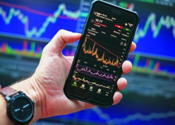 A hand holding a smartphone displaying one of the best stock trading apps, teeming with various charts and data. In the background, multiple monitors show colorful stock market graphs and candlestick charts. The person is wearing a watch on their wrist. | 5StarsStocks
