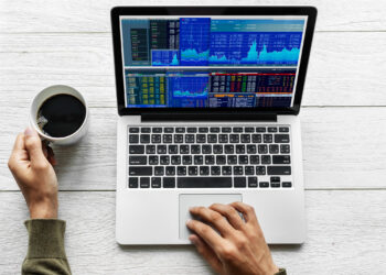 A person holds a cup of coffee while using a laptop displaying multiple financial charts and data on a wooden table. The screen shows various graphs and tables in blue and orange against a silver laptop body. | 5StarsStocks
