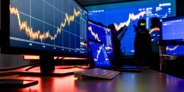 A financial trading room with multiple screens displaying dynamic stock charts. The foreground showcases detailed graphs, highlighting intricate trading strategies, while the background features a large screen and silhouetted figures analyzing data. | 5StarsStocks