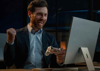 Man in a suit smiles and raises his fist in excitement while holding a game controller in front of a computer.