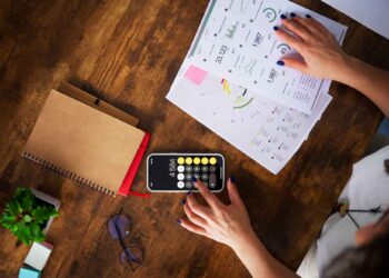 Person using a calculator on a smartphone with financial charts, notebook, glasses, and a plant on a wooden table.
