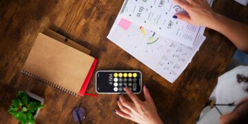 Person using a calculator on a smartphone with financial charts, notebook, glasses, and a plant on a wooden table.