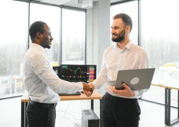 Two men in an office shake hands; one holds a laptop, stock charts are visible on a monitor behind them.