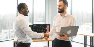 Two men in an office shake hands; one holds a laptop, stock charts are visible on a monitor behind them.