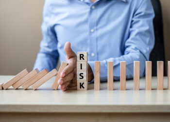 A person in a blue shirt stops falling wooden blocks with their hand. In front of their hand, vertical blocks spell "RISK," symbolizing preventing risk or stopping a chain reaction. | 5StarsStocks