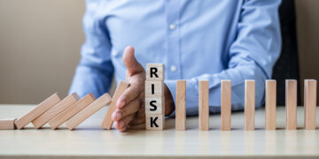 A person in a blue shirt stops falling wooden blocks with their hand. In front of their hand, vertical blocks spell "RISK," symbolizing preventing risk or stopping a chain reaction. | 5StarsStocks