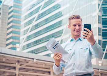 A smiling man in a light blue shirt holds a folded business newspaper and looks at his smartphone, standing outdoors in front of modern glass office buildings. | 5StarsStocks
