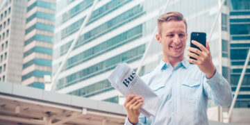 A smiling man in a light blue shirt holds a folded business newspaper and looks at his smartphone, standing outdoors in front of modern glass office buildings. | 5StarsStocks