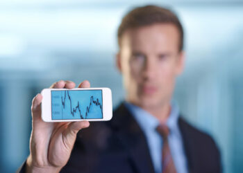 A man in a suit holds a smartphone displaying a fluctuating stock chart with numbers between 1300 and 1400; his face is out of focus in the background. | 5StarsStocks