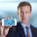 A man in a suit holds a smartphone displaying a fluctuating stock chart with numbers between 1300 and 1400; his face is out of focus in the background. | 5StarsStocks