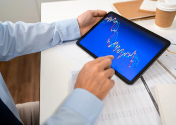 A person in a blue shirt holds a tablet displaying a colorful financial chart with fluctuating lines, at a desk with documents, a notepad, and a coffee cup. | 5StarsStocks