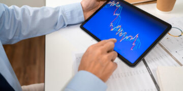 A person in a blue shirt holds a tablet displaying a colorful financial chart with fluctuating lines, at a desk with documents, a notepad, and a coffee cup. | 5StarsStocks