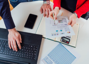 Three people working at a desk with a laptop, smartphone, calculator, documents, and charts. One person points at a printed report with graphs while another types on the laptop. | 5StarsStocks