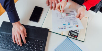 Three people working at a desk with a laptop, smartphone, calculator, documents, and charts. One person points at a printed report with graphs while another types on the laptop. | 5StarsStocks
