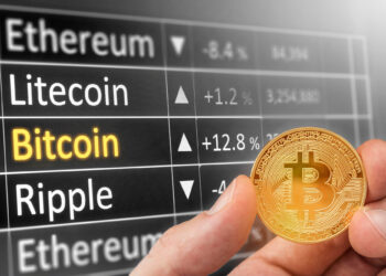 A hand holds a physical Bitcoin coin in front of a digital board displaying cryptocurrency names—Ethereum, Litecoin, Bitcoin, Ripple—and their percentage changes. Bitcoin is highlighted in yellow. | 5StarsStocks