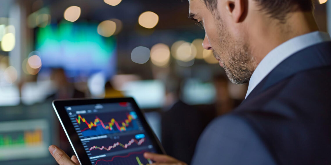 A man in a suit uses a tablet displaying colorful financial charts and graphs in a modern office with blurred lights and monitors in the background. | 5StarsStocks