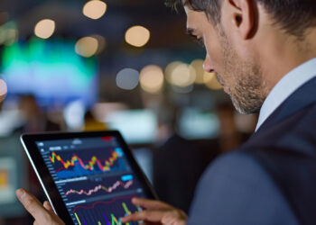 A man in a suit uses a tablet displaying colorful financial charts and graphs in a modern office with blurred lights and monitors in the background. | 5StarsStocks
