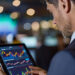 A man in a suit uses a tablet displaying colorful financial charts and graphs in a modern office with blurred lights and monitors in the background. | 5StarsStocks