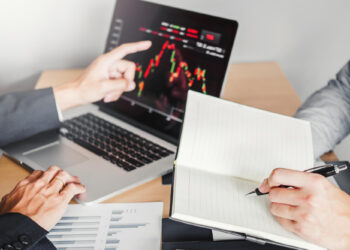 Two people in business attire discuss a laptop screen displaying a financial chart. One points at the screen, while the other writes in a notebook. Papers with graphs are on the table. | 5StarsStocks