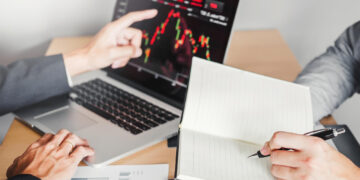 Two people in business attire discuss a laptop screen displaying a financial chart. One points at the screen, while the other writes in a notebook. Papers with graphs are on the table. | 5StarsStocks