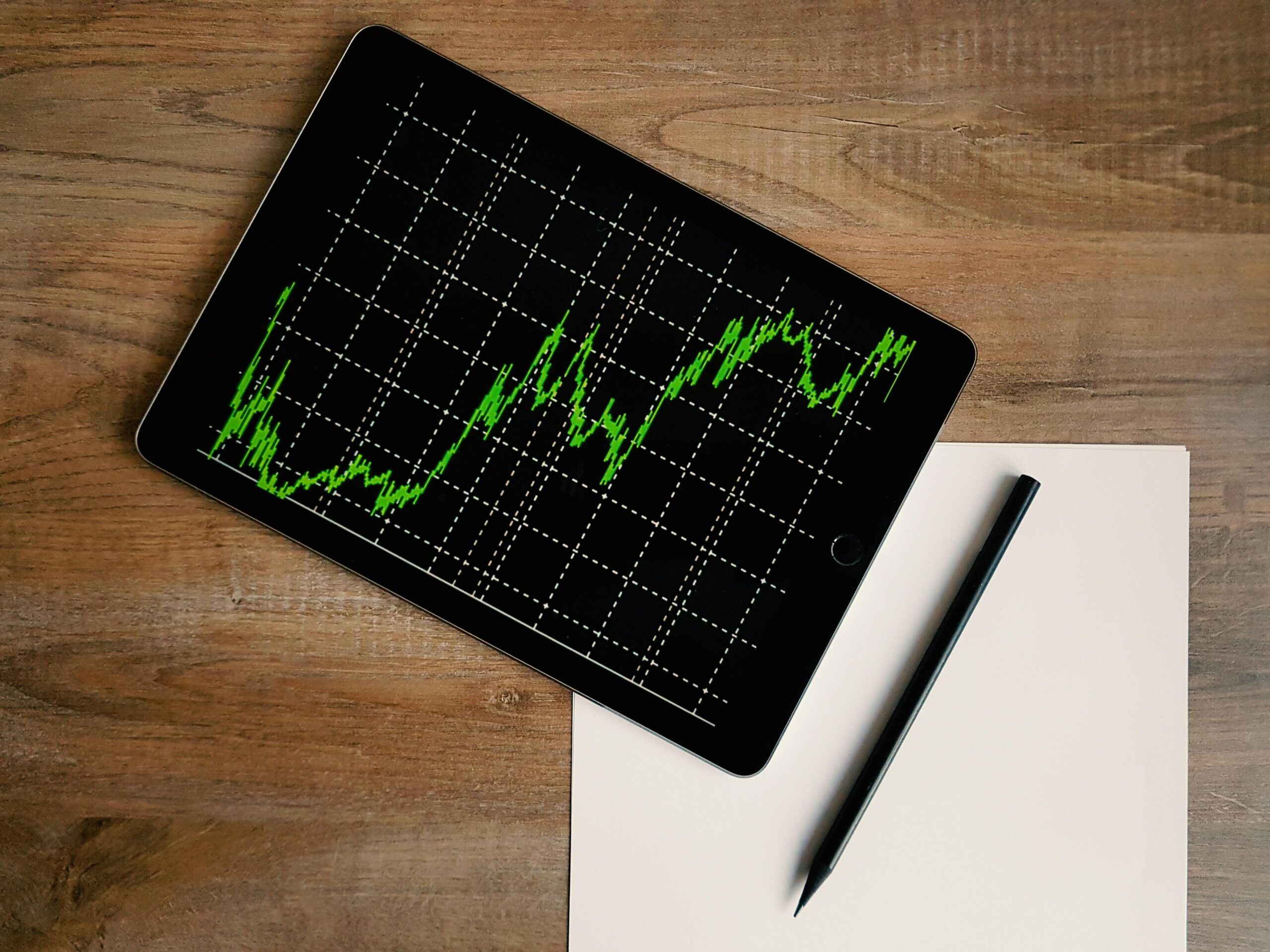 A tablet displaying a green fluctuating line graph related to investing rests on a wooden desk next to a blank sheet of white paper and a black pen. | 5StarsStocks