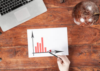 A hand points to a bar graph with red bars on a sheet of paper, placed on a wooden table next to a laptop and a small globe. | 5StarsStocks