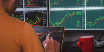 A person in an orange shirt analyzes stock charts on a tablet, surrounded by large monitors displaying various financial graphs; a red coffee mug and computer mouse are on the desk. | 5StarsStocks