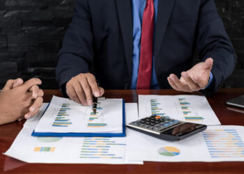 Two people in business attire sit at a desk covered with financial charts, graphs, a calculator, and a laptop. One person gestures while discussing the documents, indicating a business meeting or financial review. | 5StarsStocks