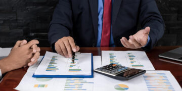 Two people in business attire sit at a desk covered with financial charts, graphs, a calculator, and a laptop. One person gestures while discussing the documents, indicating a business meeting or financial review. | 5StarsStocks