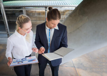 Two business professionals stand outdoors, reviewing documents with charts and notes. The woman holds a clipboard with colorful graphs, while the man looks at a notebook. They appear to be discussing business data. | 5StarsStocks