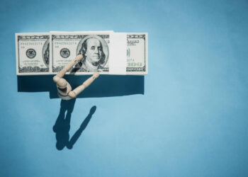 A wooden mannequin arm reaches toward a row of three hundred-dollar bills on a flat blue background, casting a shadow below. | 5StarsStocks